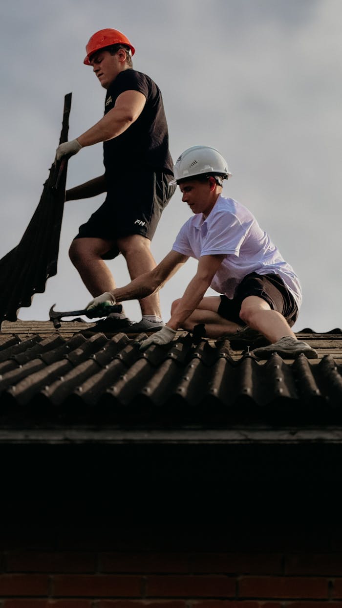Two construction workers fixing roof tiles, emphasizing teamwork and safety with helmets.